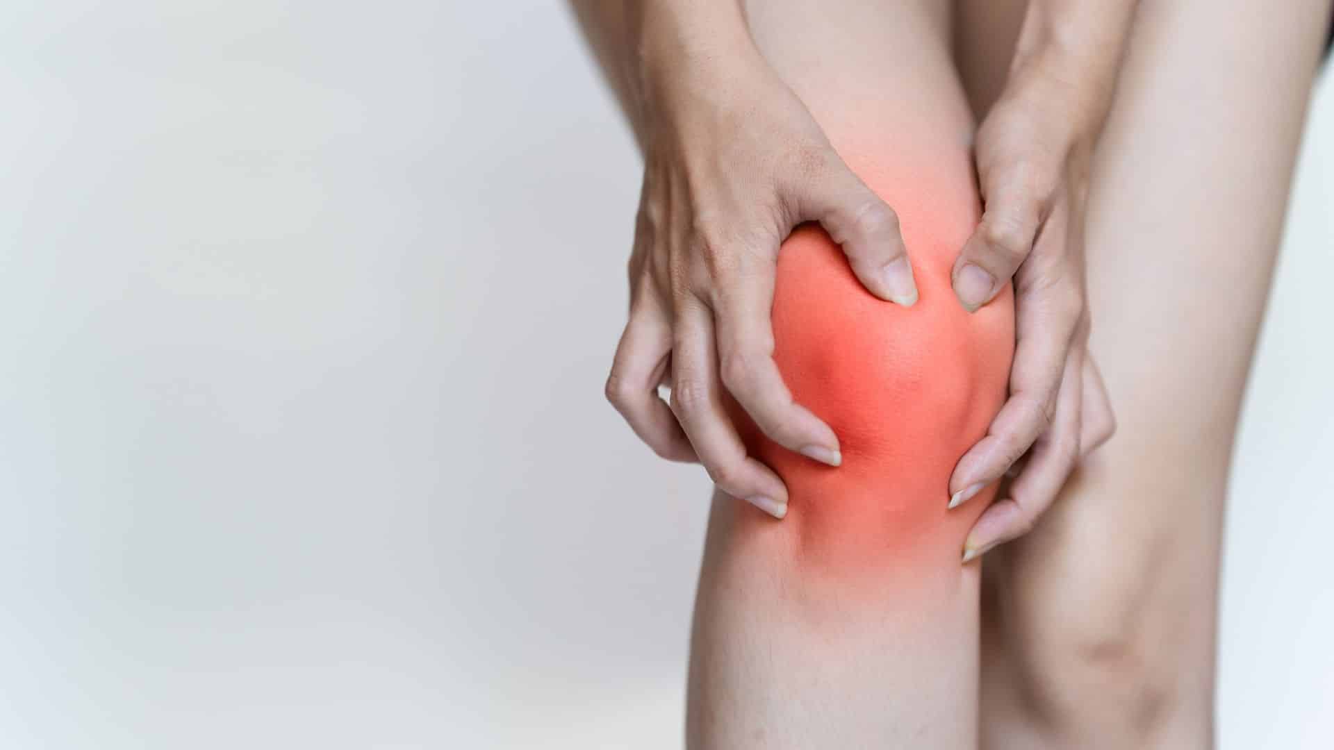 A person is clutching their knee, which is visibly inflamed and red, possibly indicating pain or injury. The background is plain and out of focus, emphasizing the discomfort in the knee area.