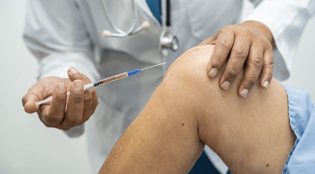 A healthcare professional, wearing a white coat and stethoscope, prepares to administer an injection into a patient's bent knee. The patient is wearing a blue garment, and both the hands of the professional and the syringe filled with medication are in focus.