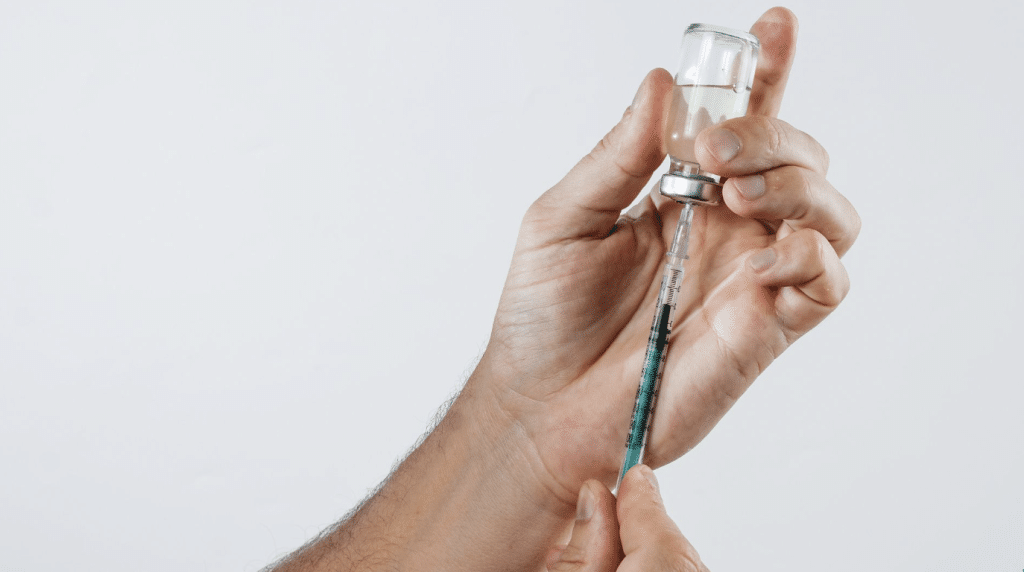 Hands holding a syringe that is being filled with a clear liquid from a vial, against a plain white background.