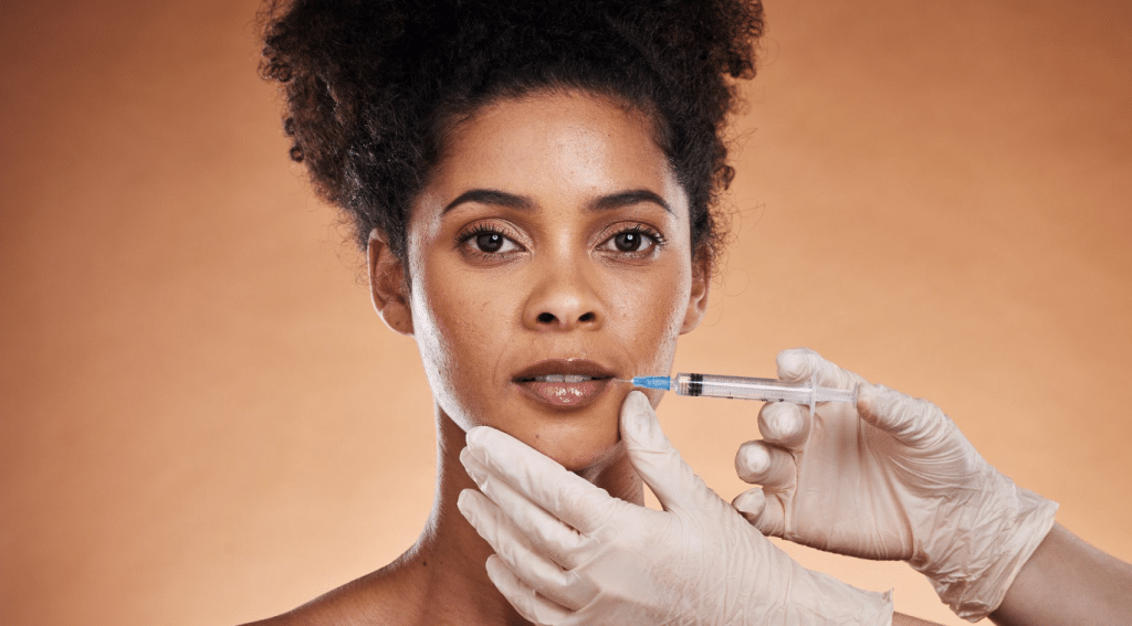 A close-up of a person receiving a cosmetic injection in the lips. They have a calm expression. Gloved hands hold the syringe and gently touch their chin. The background is a soft orange-brown gradient.