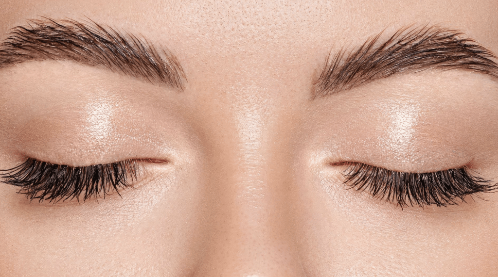 Close-up image of a person's face focusing on their closed eyes. The person has smooth skin, well-defined eyebrows, and long, thick eyelashes. The eyelids have a slight shimmer, suggesting the application of a light, glowy makeup.