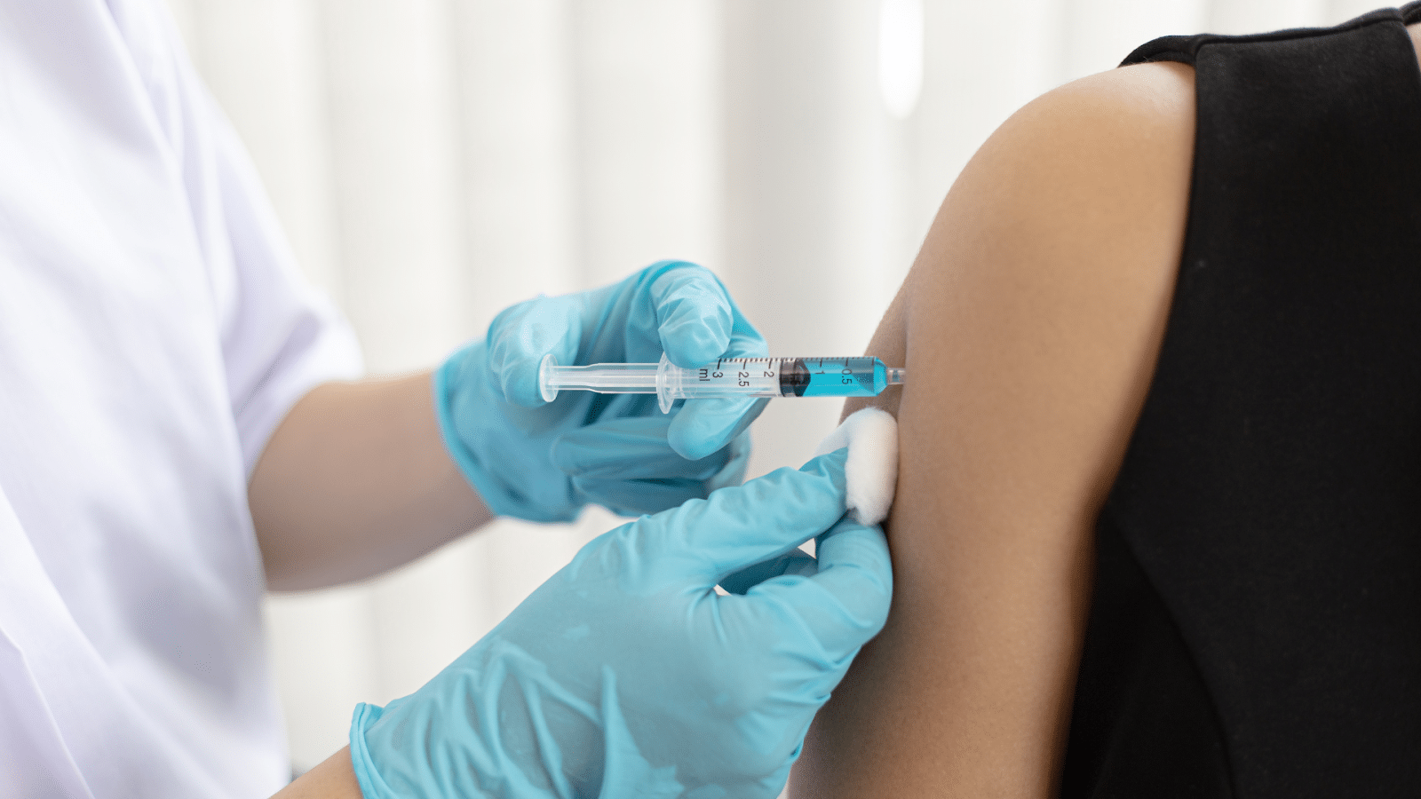 A healthcare professional wearing blue gloves administers a vaccine using a syringe to a person's upper arm. The arm is being held steady with a cotton ball. The person receiving the vaccine is wearing a black sleeveless top.