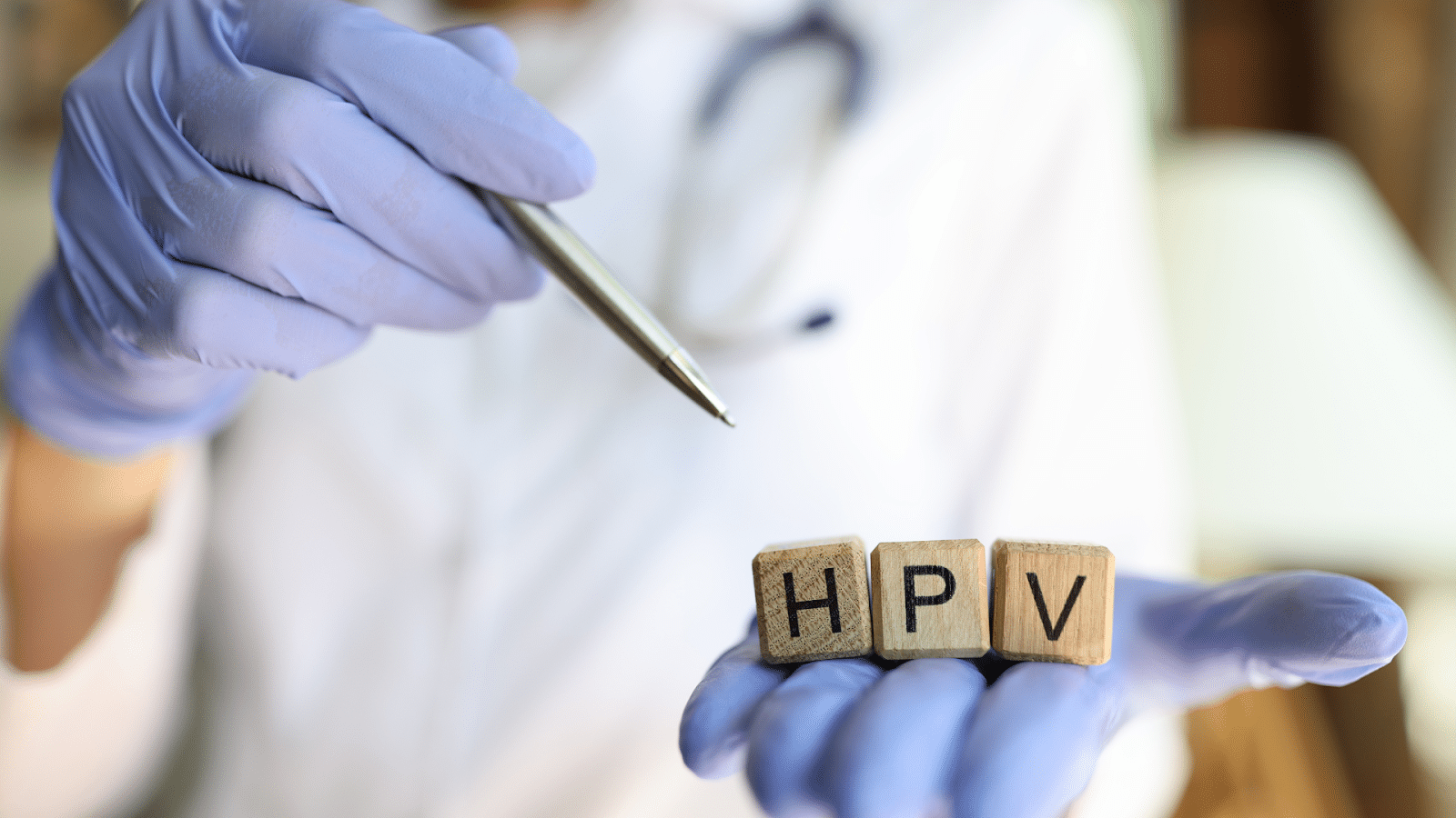 A person wearing blue gloves holds three wooden blocks with the letters "H", "P", and "V". They are pointing at the blocks with a pen. The background is blurred, focusing attention on the blocks and the person's hand.