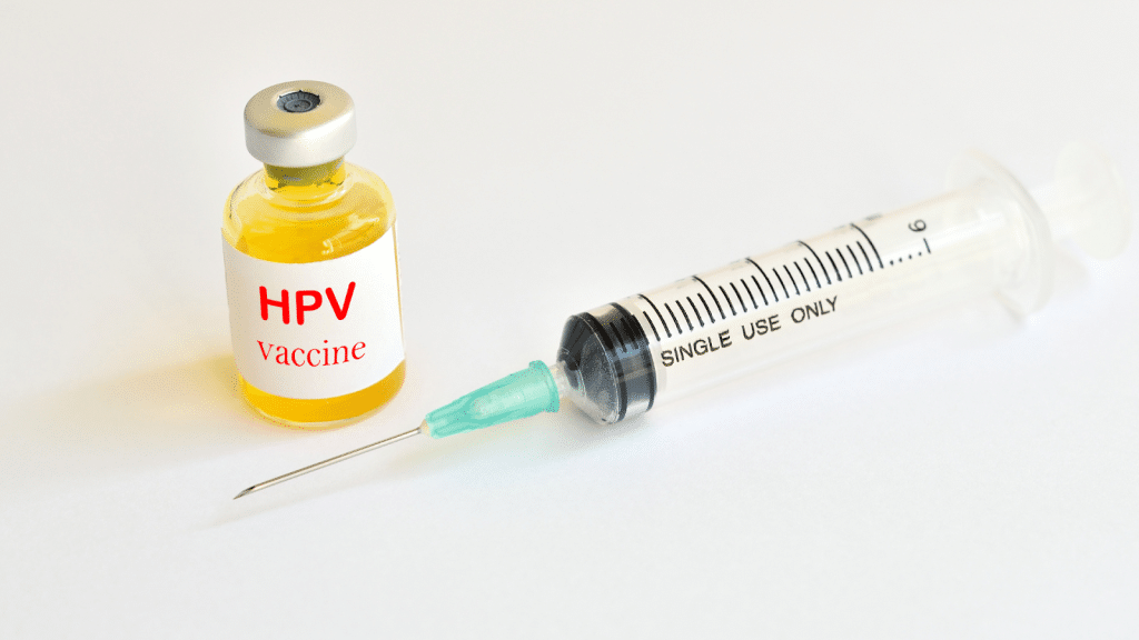 A vial labeled "HPV vaccine" containing yellow liquid is placed next to a syringe with a needle, marked "Single Use Only." The items are on a white background, suggesting a medical or clinical setting.