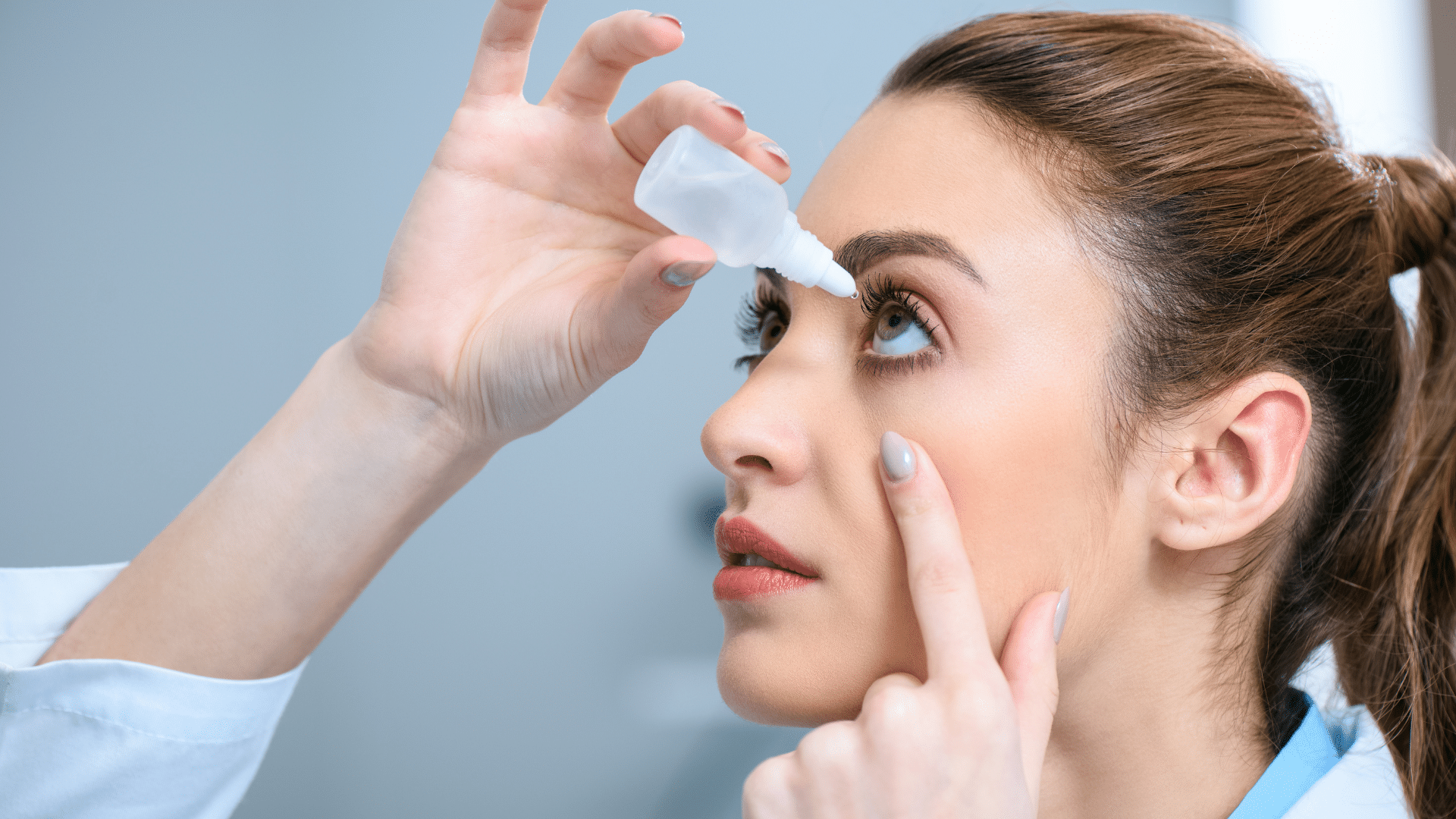 A close-up of a person with their head tilted back, looking up as they prepare to apply eye drops with a small bottle held in one hand while using the other hand to hold their lower eyelid down. The background is blurred.