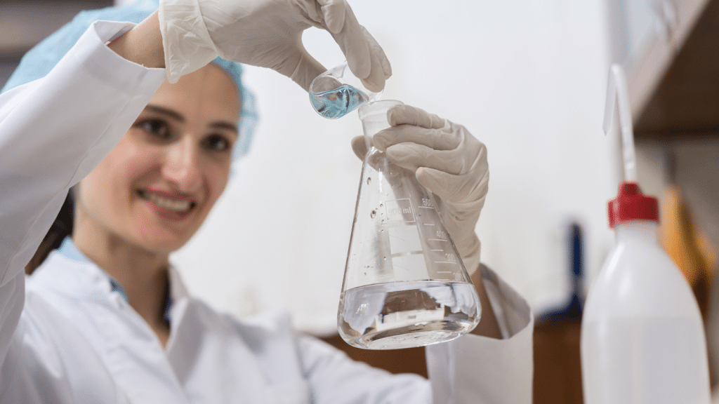 A woman in a lab coat and protective gear is smiling as she pours a blue liquid from a small container into a conical flask filled with a clear liquid in a laboratory setting.