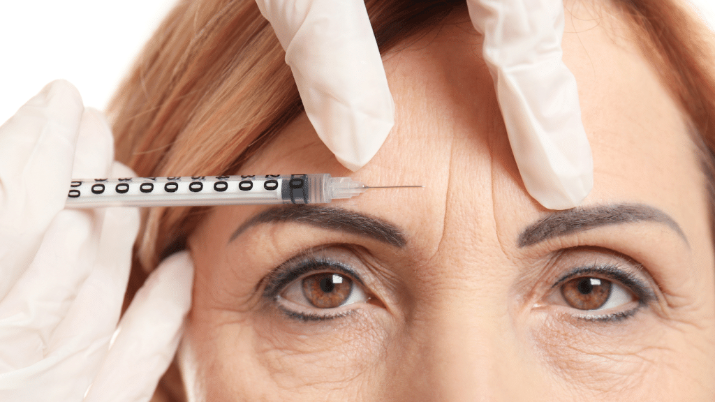 A close-up of a woman’s face as she receives an injection between her eyebrows. She has brown eyes and light brown hair, and wears light makeup. The person administering the injection is wearing white gloves.