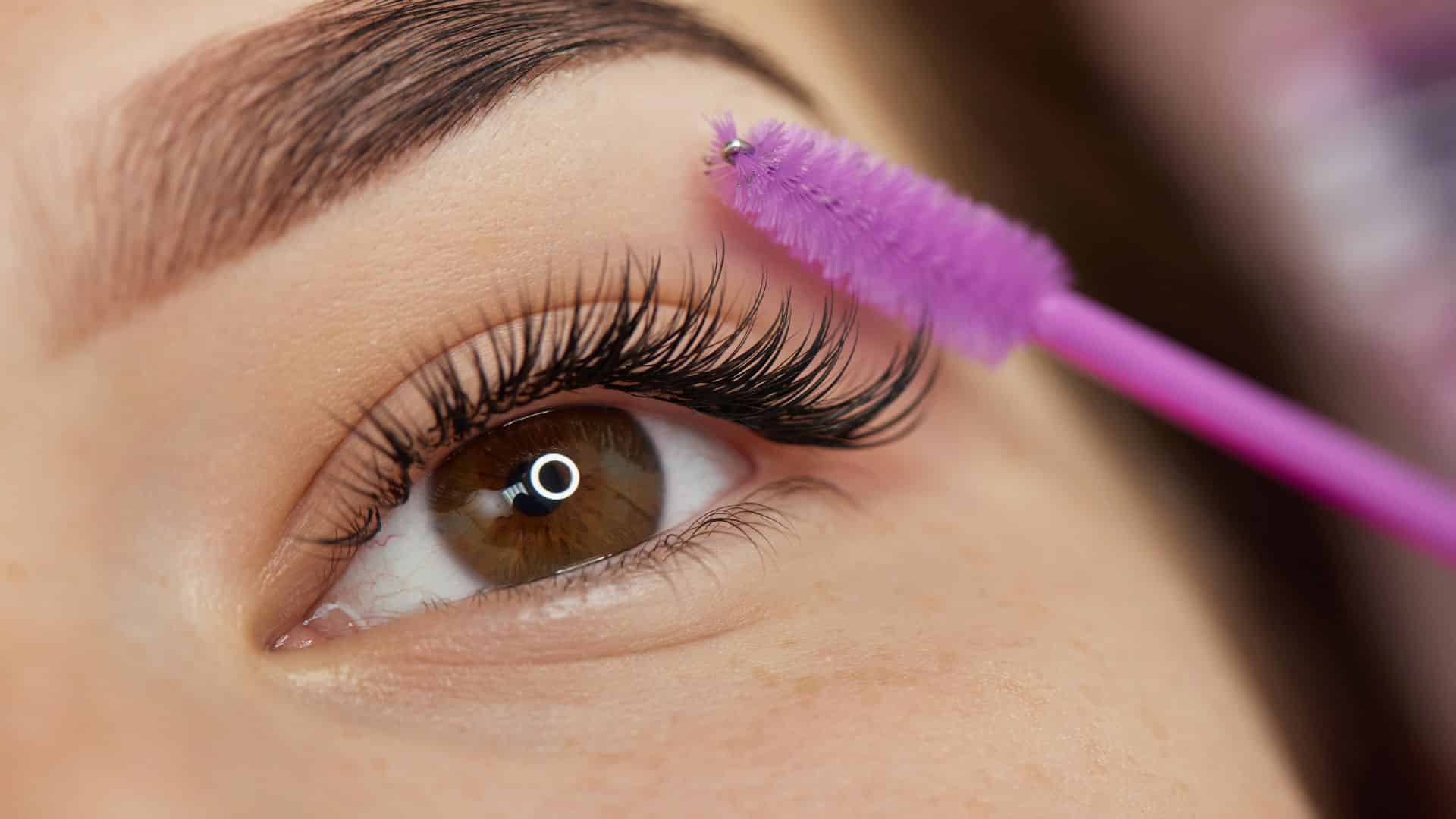 Close-up of an eye with long, dark eyelashes being brushed with a purple eyelash brush. The brow above the eye is neatly groomed, and the skin around the eye is clear. The scene focuses on the makeup application process.