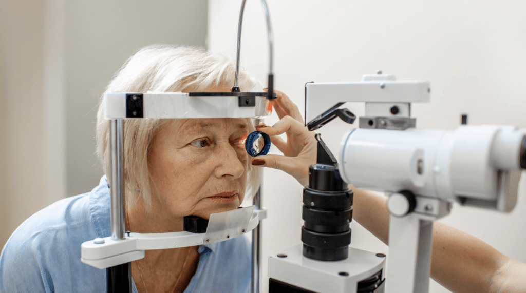 An elderly woman undergoing an eye examination with a slit lamp at an optometrist's office. The optometrist is holding a lens in front of her left eye while the woman rests her chin on the machine. The background is a plain, light-colored wall.