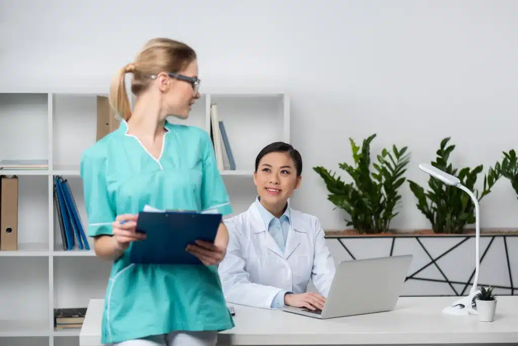 A nurse in scrubs, holding a clipboard, stands next to a seated doctor wearing a white coat and working on a laptop. They are in a bright, modern office with a bookshelf, folders, and green plants in the background.