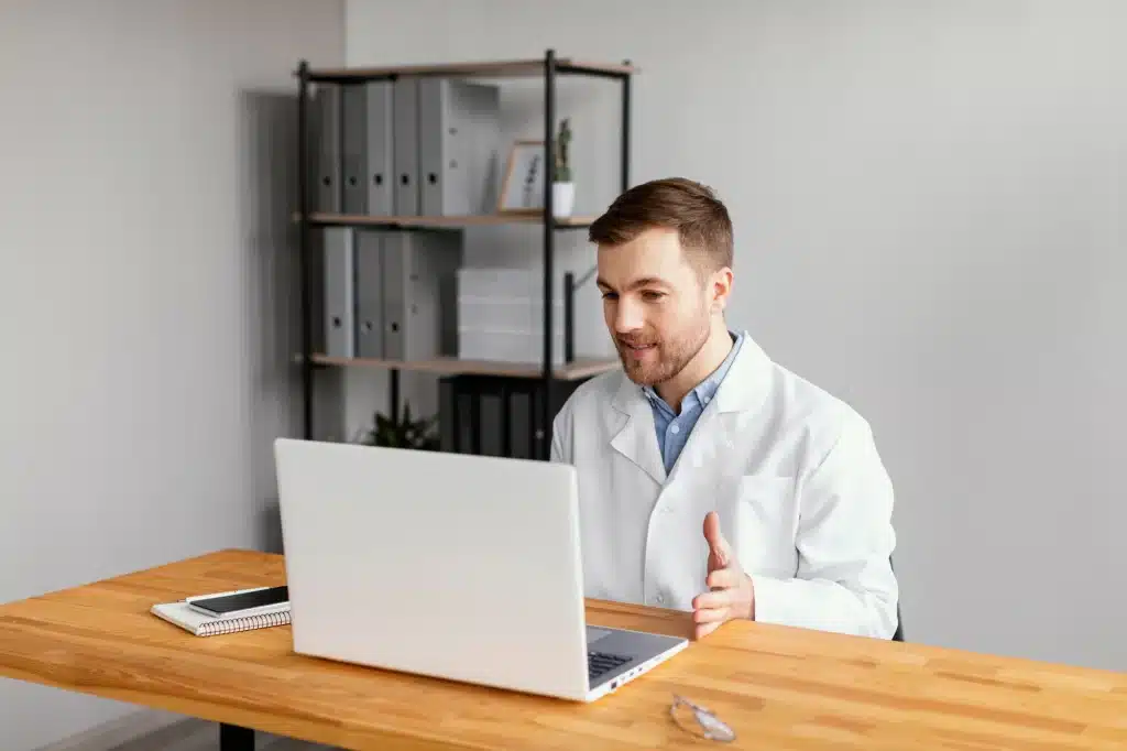 Young doctor with a white labcoat, looking at his laptop. He is sitting at the desk, inside an office