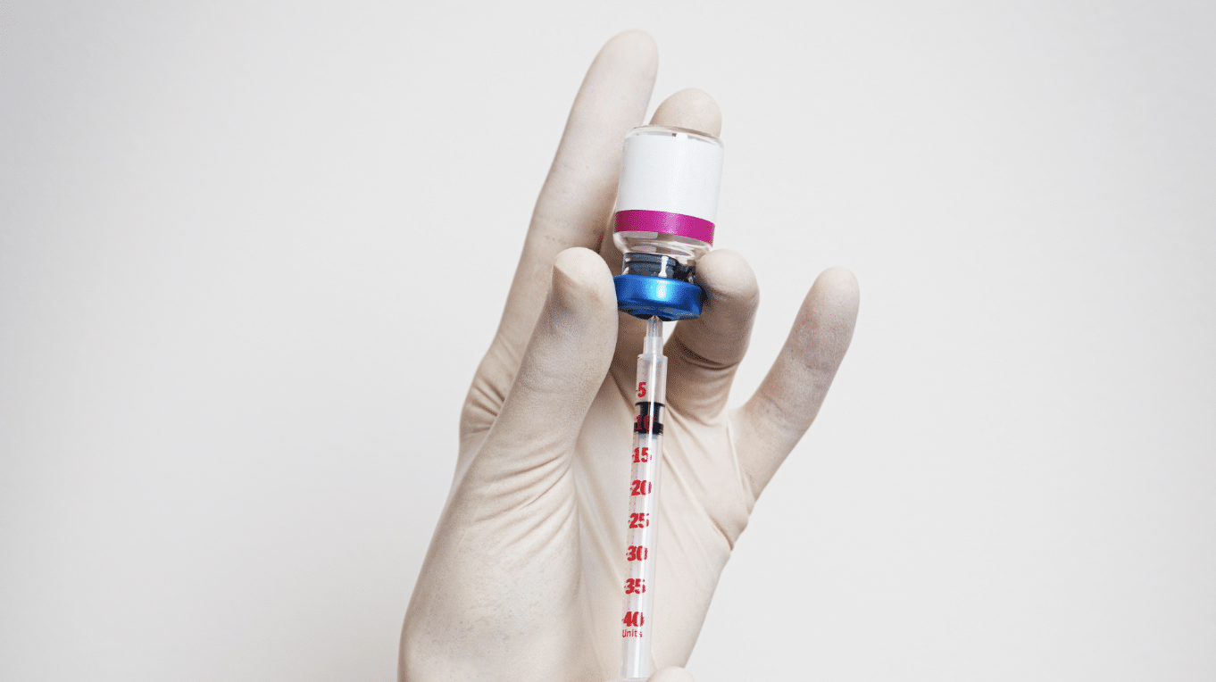 A gloved hand holds a vial and a syringe that is drawing liquid from the vial. The background is plain white.