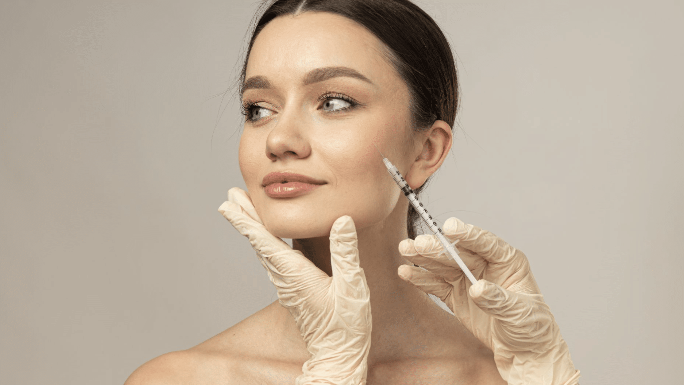 A woman with brunette hair pulled back is receiving an injection in her cheek from a medical professional wearing gloves. The woman has a serene expression and the background is plain, focusing attention on the procedure.