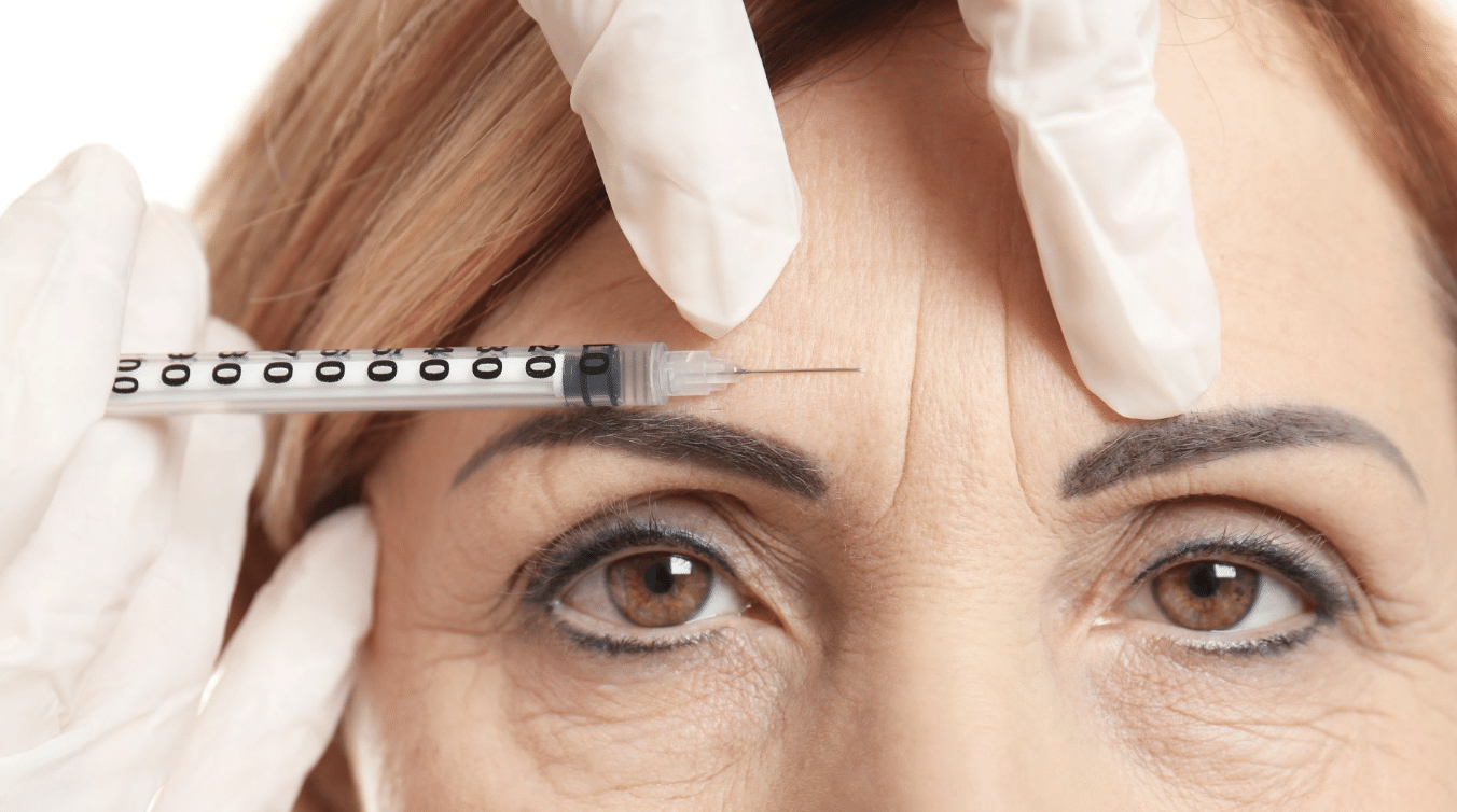 Close-up of a woman receiving a cosmetic injection to her forehead. The person administering the injection is wearing white gloves and holding a syringe with the needle close to the woman's forehead. The woman's eyes are open, looking straight ahead.