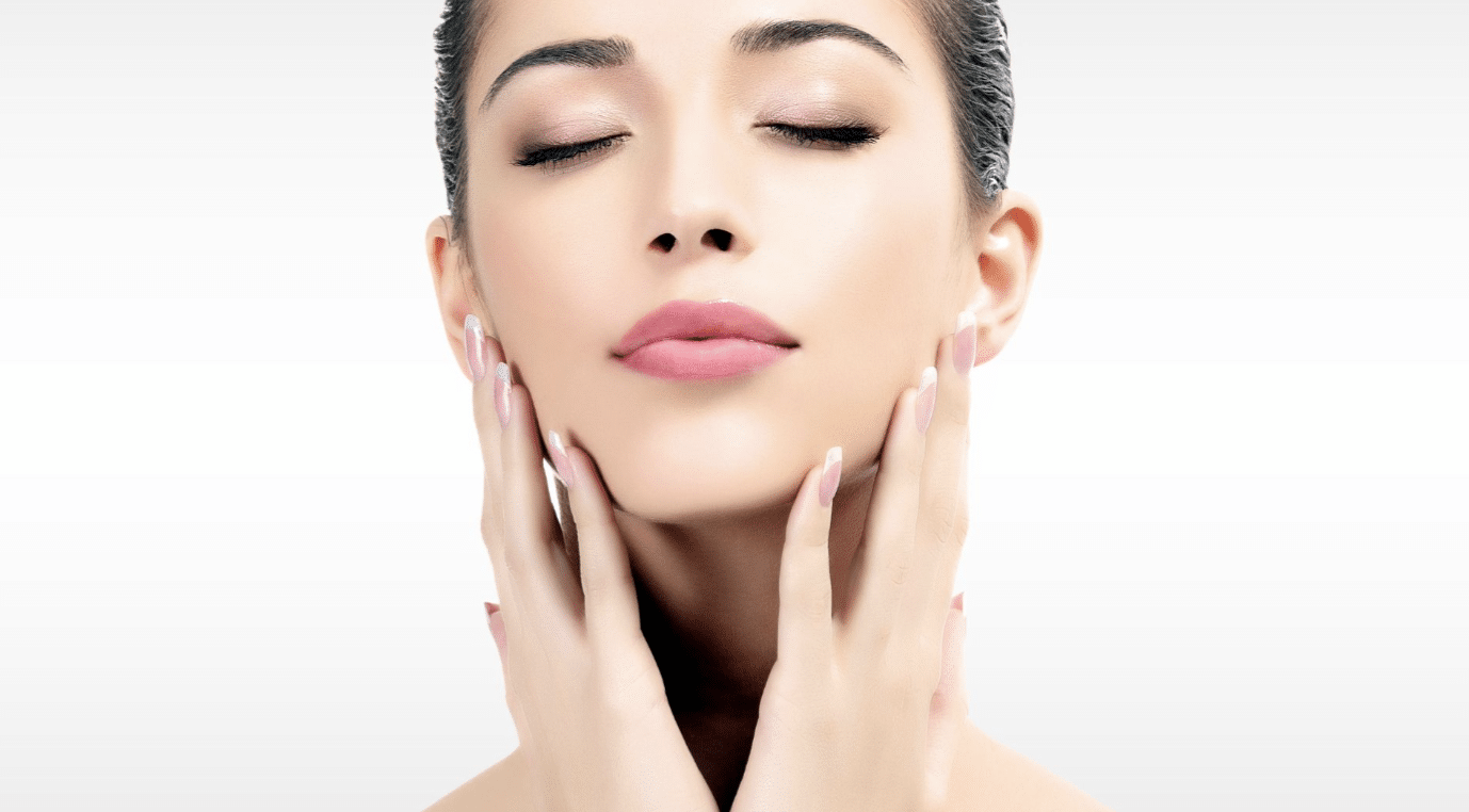 A woman with smooth skin and closed eyes gently touching her cheeks with both hands. She has light makeup with pink lipstick and a serene expression. The background is a gradient of light grey to white.