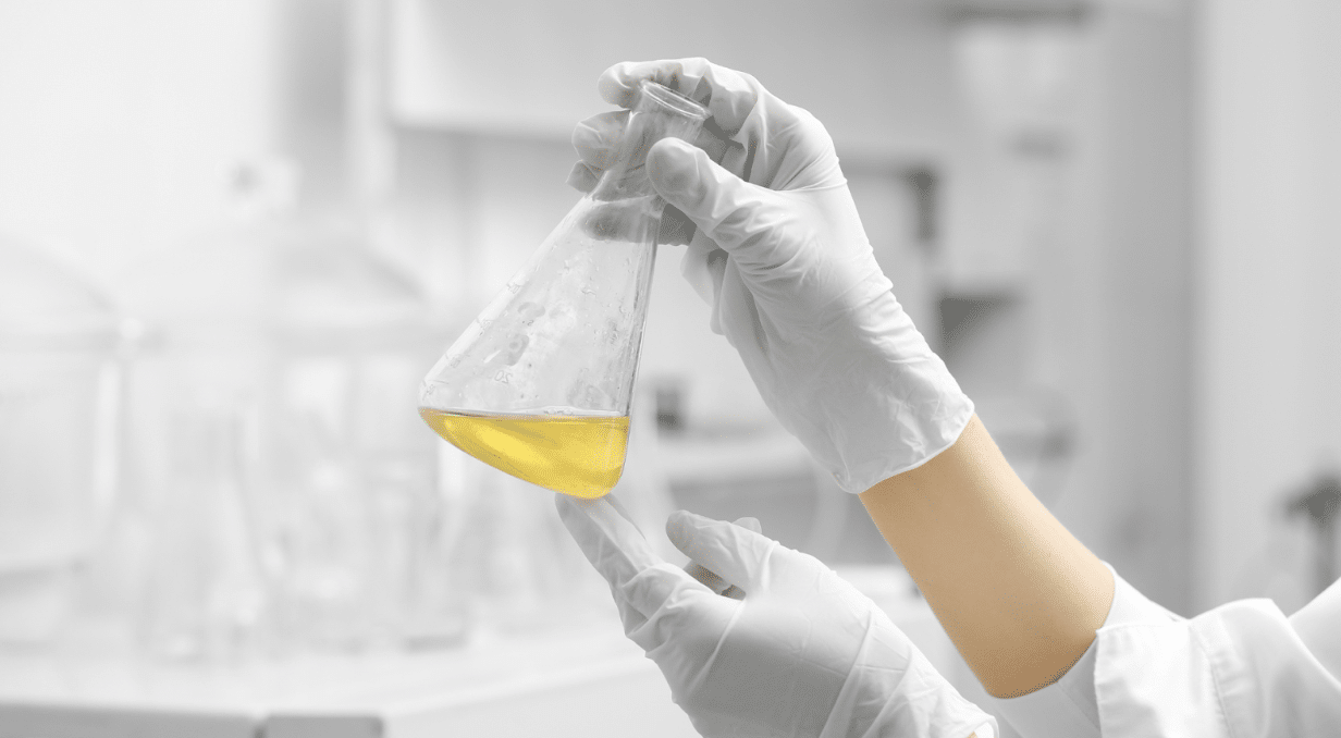 A gloved person holds up an Erlenmeyer flask containing a yellow liquid in a laboratory setting. Blurred lab equipment can be seen in the background.