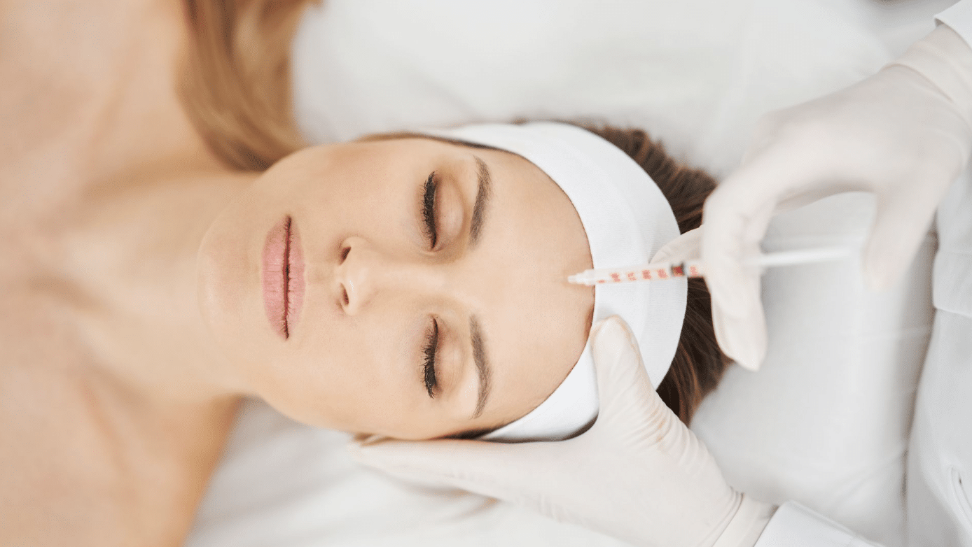 A person wearing a white headband lies with eyes closed while a gloved hand injects a syringe into their forehead.