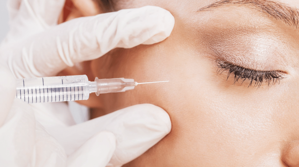 Close-up of a person receiving a cosmetic injection near their right eye. The person's eyes are closed, and a gloved hand is holding a syringe containing a clear liquid, preparing to administer the injection. The skin appears smooth and well-groomed.