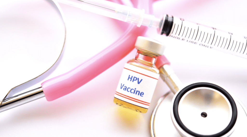 A syringe, stethoscope, and a small vial labeled "HPV Vaccine" are placed on a white surface. The stethoscope has pink tubing and a silver chest piece.