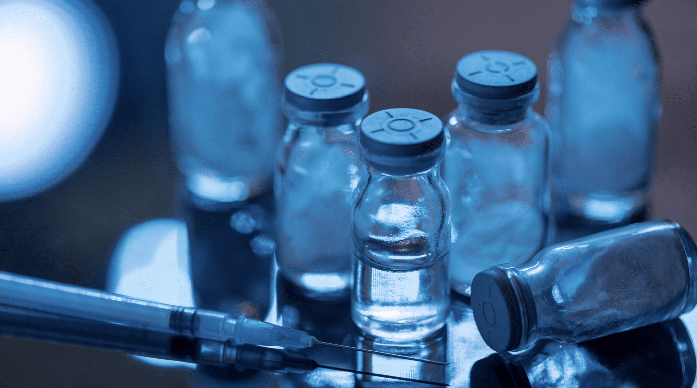 Close-up of several transparent glass vials filled with a clear liquid, some sealed with metal caps, lying on a reflective surface. A syringe with a needle is positioned in the foreground. The image has a blue tint, giving it a clinical and sterile feel.