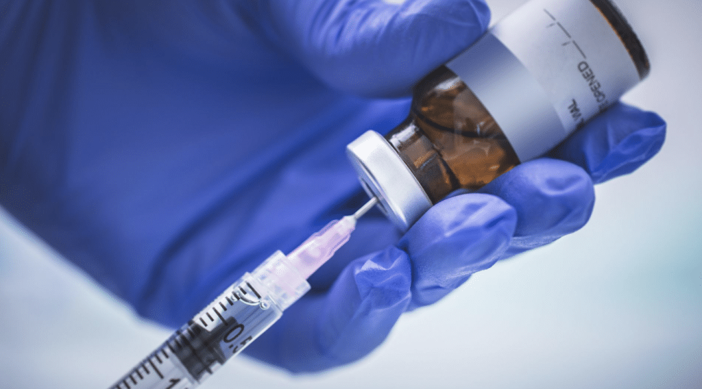 Close-up of a gloved hand holding a brown vial and drawing liquid from it into a syringe. The needle is inserted into the vial’s rubber stopper, preparing to extract or inject the fluid inside. The scene depicts a medical or laboratory setting.