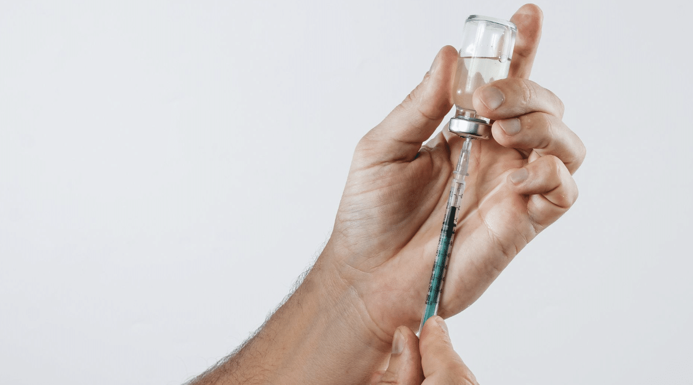 Close-up of a person's hands holding a vial and filling a syringe with liquid. The person is using one hand to steady the vial while the other hand draws the liquid into the syringe, which has measurements marked on it. The background is a plain white.