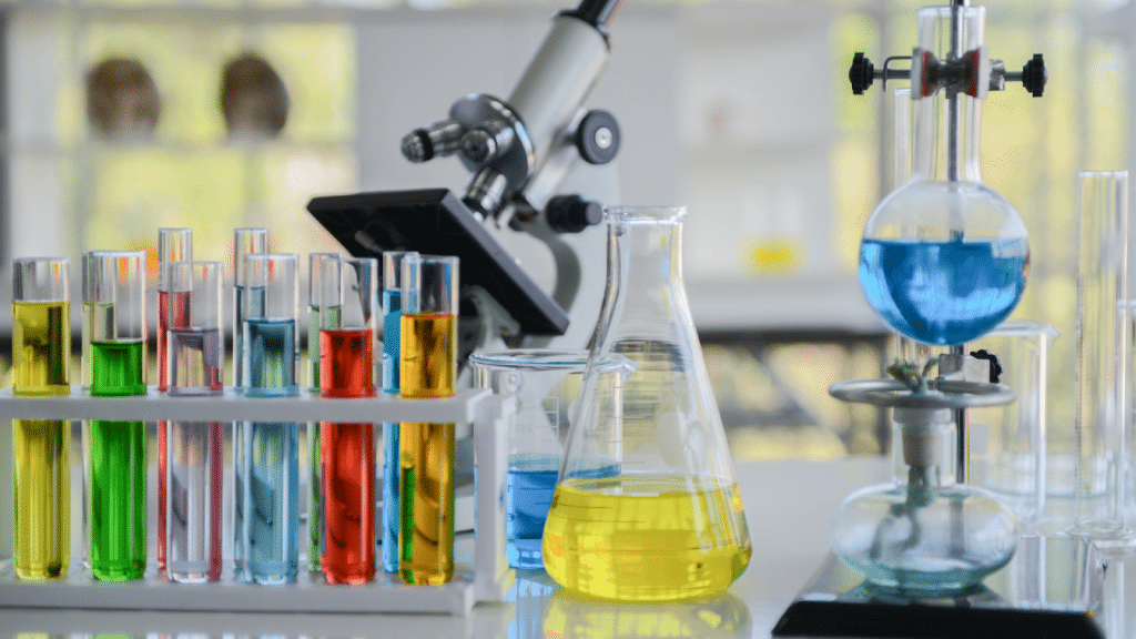 A lab bench displaying various scientific equipment. There are test tubes filled with colorful liquids in a rack, a yellow liquid in a beaker, blue liquid in a round-bottom flask on a stand, and a microscope in the background.