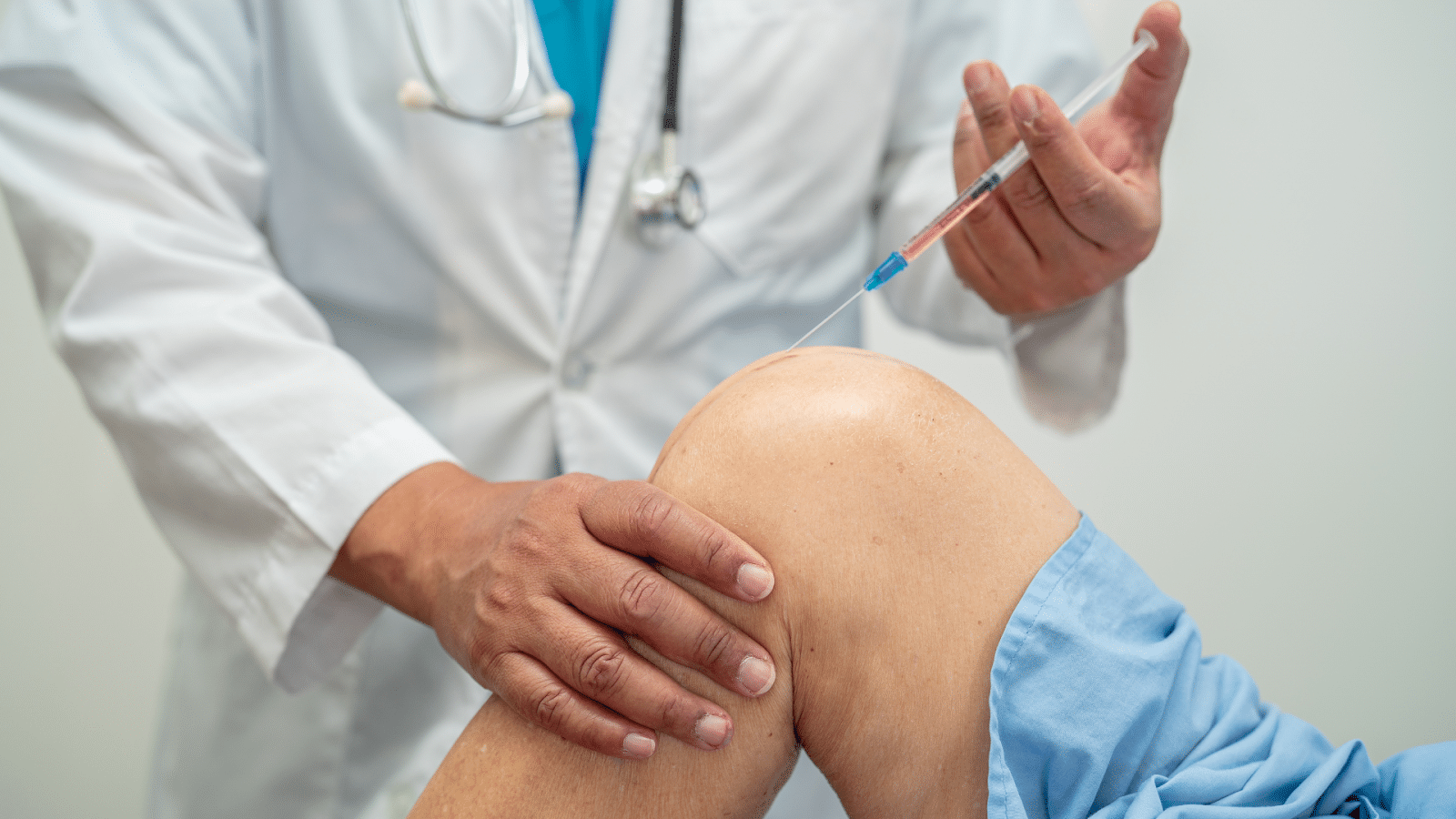 A healthcare professional in a white coat is performing a procedure on a patient's knee, holding a syringe with a clear liquid. The patient is seated with their knee bent and is wearing a blue medical gown.