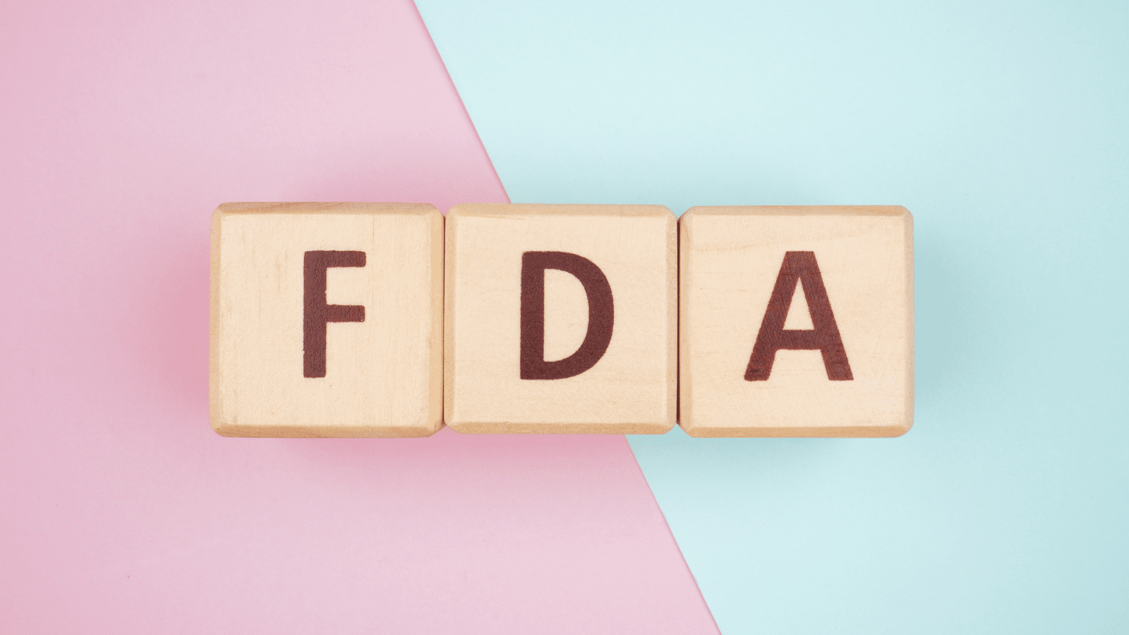 Wooden blocks with the letters "FDA" are displayed against a background divided diagonally into two colors: pink on the left and light blue on the right.