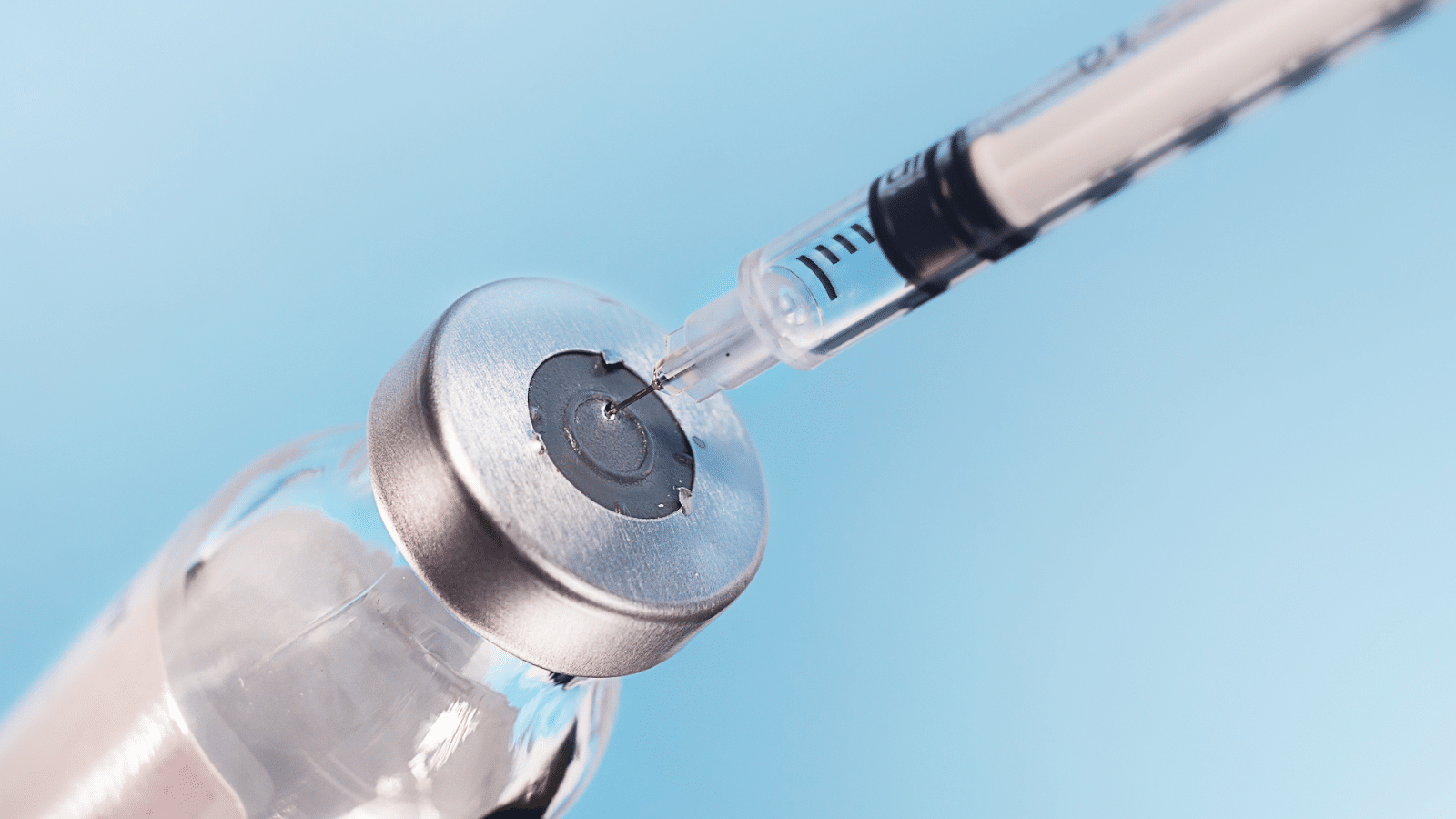 Close-up of a syringe with a needle penetrating the rubber stopper of a glass vial containing a liquid. The background is blurred and light blue in color.