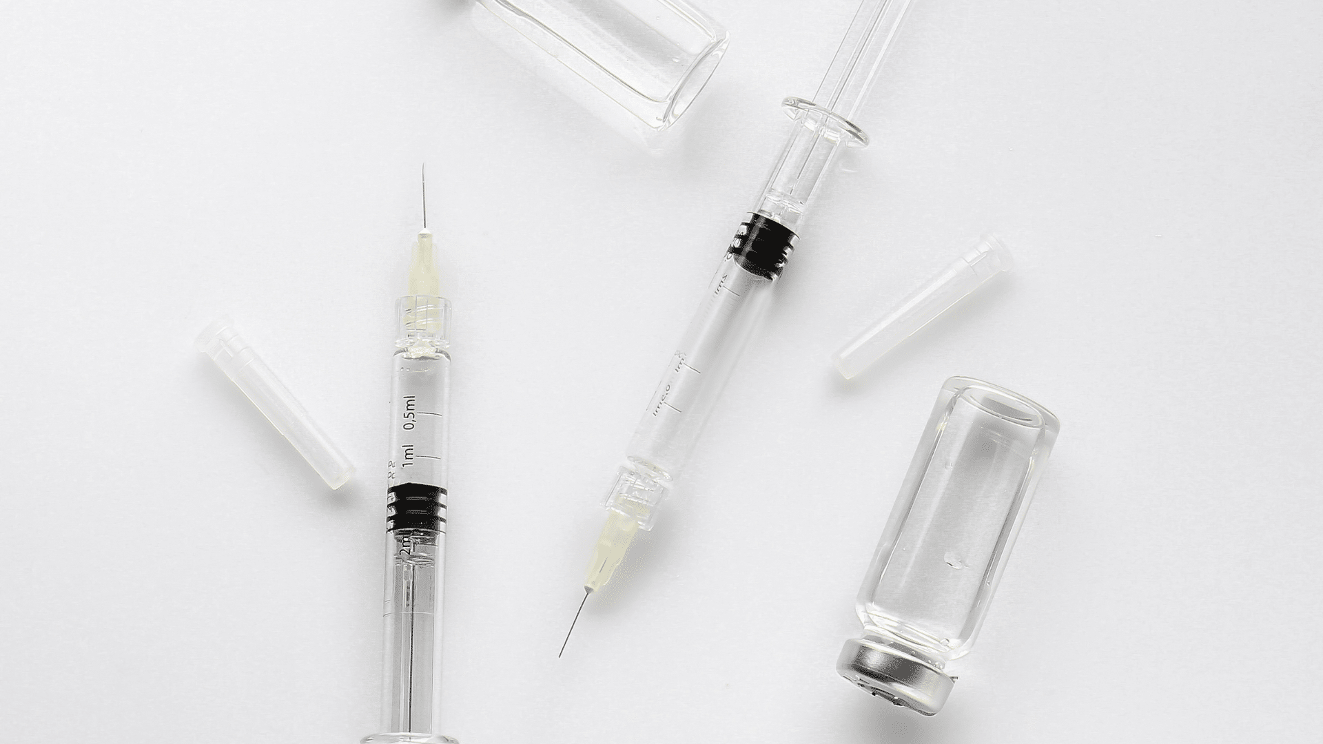 Two syringes and one vaccine vial are placed on a white surface. The syringes have clear barrels and black plunger seals, and the needles are exposed. The vial is transparent and sealed with a metal cap. Some syringe caps are also visible nearby.