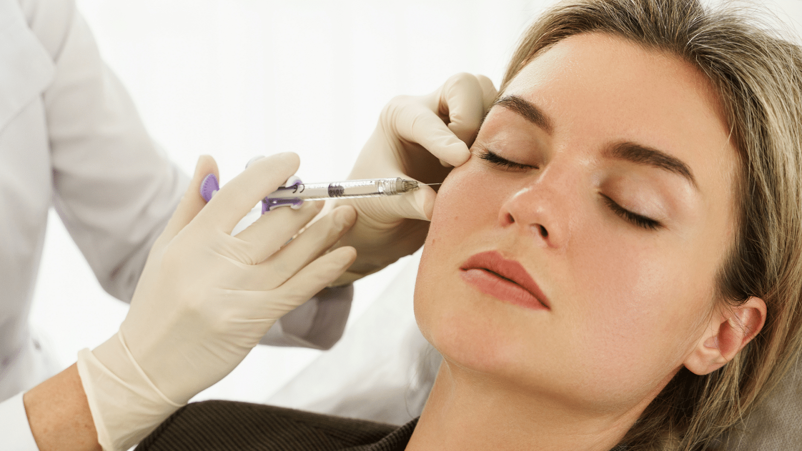 A woman with her eyes closed receives a cosmetic injection on her cheek from a medical professional wearing white latex gloves. She appears calm and relaxed during the procedure.