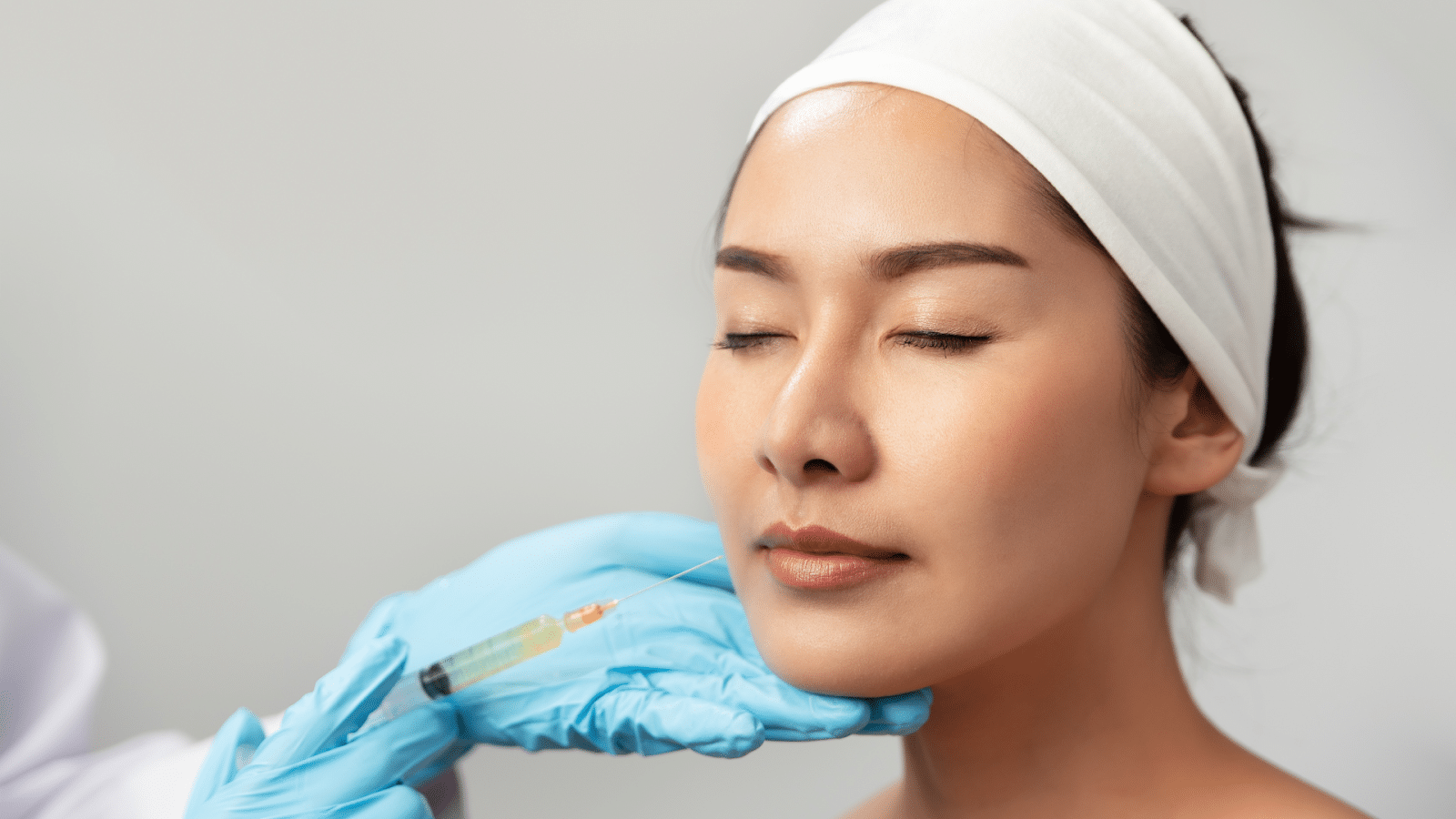 A person with a headband and closed eyes receiving a cosmetic facial injection in the lower cheek or chin area from a gloved hand.