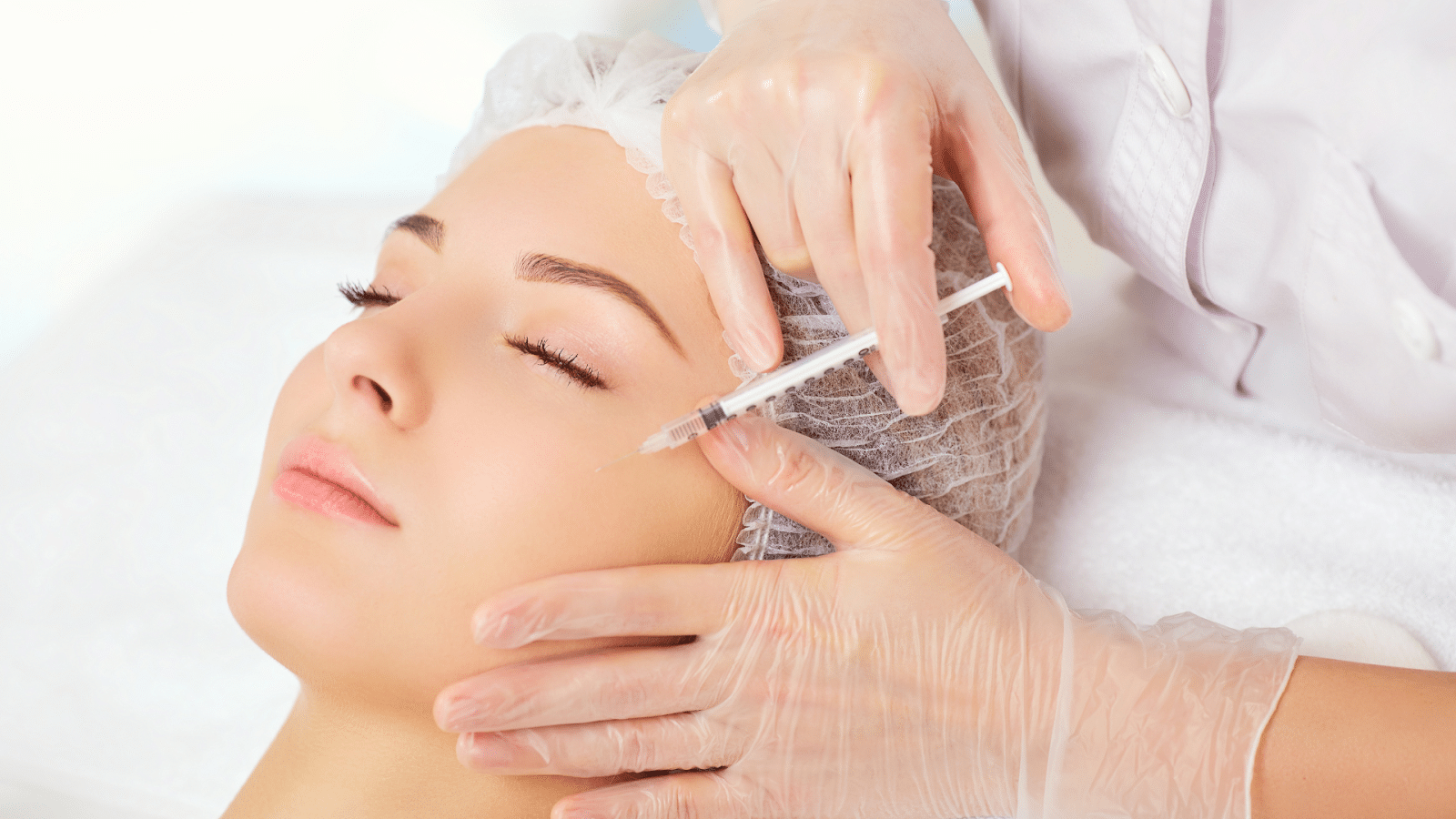 A woman with her eyes closed is receiving a facial injection from a professional wearing gloves and a white protective cap. The woman is lying down, and the professional is holding a syringe near her forehead.