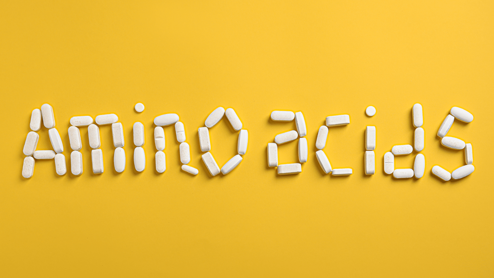 White capsules arranged to spell "Amino acids" on a bright yellow background. The capsules are uniformly shaped and placed creatively to form each letter, making the text both readable and visually appealing.