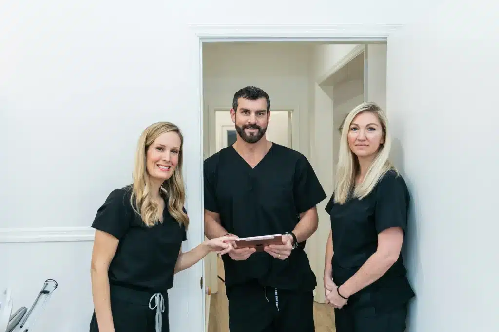 Three healthcare professionals in black scrubs stand in a white-walled hallway, smiling. One is holding a tablet. A medical device is visible on the left side, indicating a clinical setting.