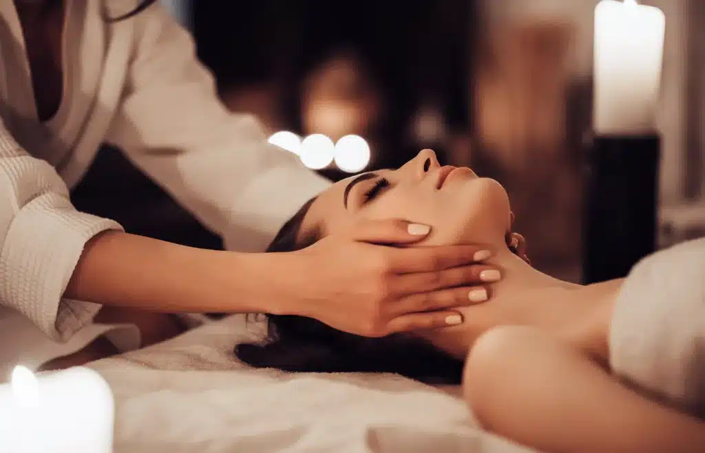 A person receiving a relaxing massage at a spa. The individual lies on a soft surface with eyes closed, while the masseuse gently holds their head. Soft lighting and lit candles create a serene ambiance.