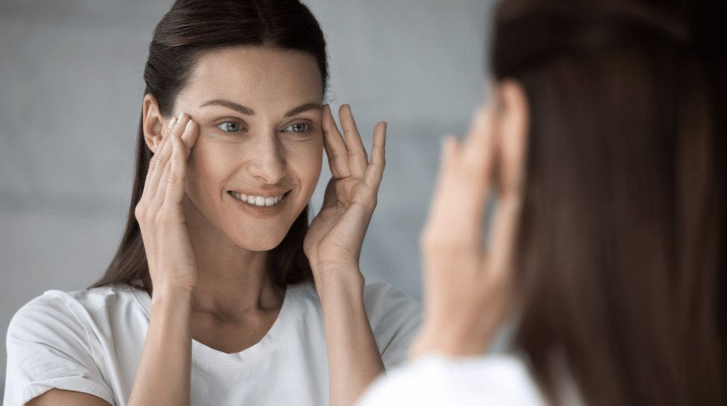 A woman with long, dark hair is smiling while touching her face with both hands, standing in front of a mirror. She is wearing a white shirt and appears to be examining her reflection with a content expression.