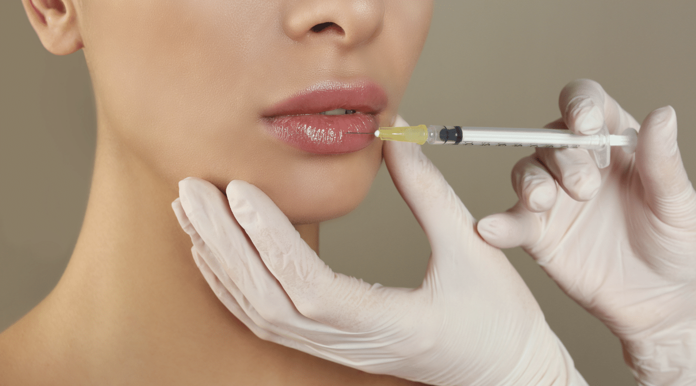 A close-up of a person receiving a injection in the lips. The person administering the shot is wearing white gloves and holding the syringe carefully. The background is blurred and neutral-toned.