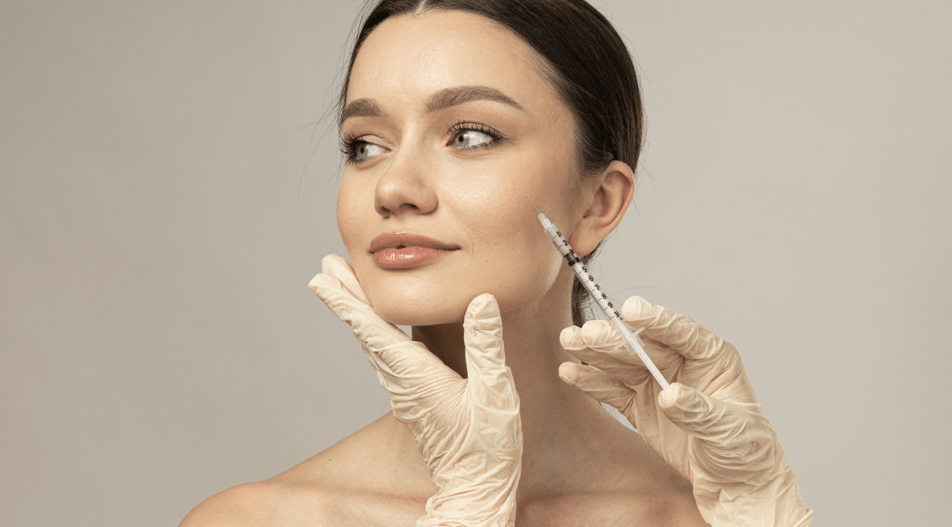 A person with smooth skin receives a beauty or medical injection on the side of their face. The individual administering the injection wears yellowish latex gloves and gently holds the client's jaw while preparing to inject with a syringe.