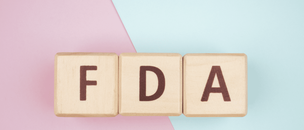 Three wooden blocks arranged in a row on a two-toned background of pink and light blue. Each block has a single letter inscribed, together spelling "FDA.