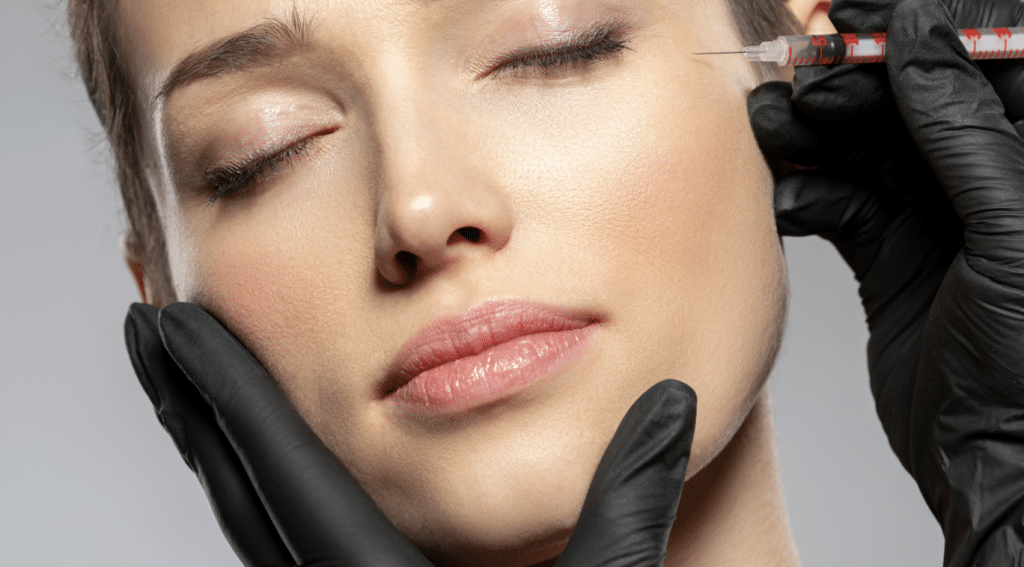 Close-up of a woman's face with closed eyes while a black-gloved hand holds her cheek and another gloved hand administers an injection near her temple. The woman appears relaxed, suggesting a cosmetic or medical procedure.