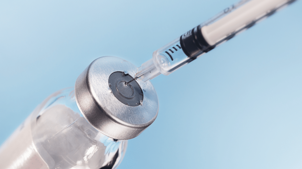 Close-up of a syringe with a needle puncturing the gray rubber seal of a clear glass vial, against a light blue background. The syringe appears to be extracting or injecting liquid into the vial.