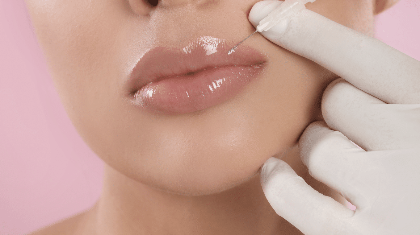 Close-up image of a person with glossy, plump lips receiving a filler injection in the upper lip. A gloved hand holds a syringe, and the background is a soft pink color.