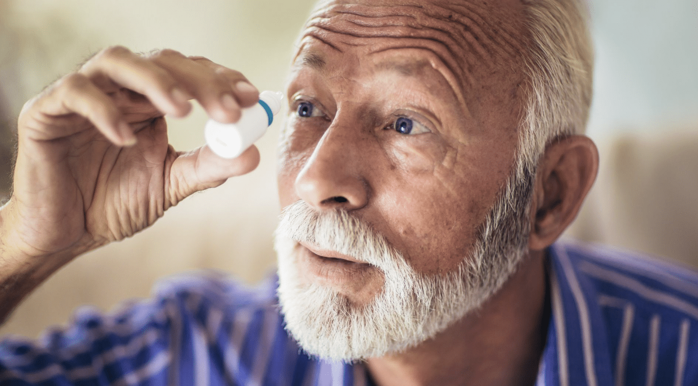 An elderly man with white hair and beard is holding an eye drop bottle close to his open eye, preparing to apply the eye drops. He is wearing a blue striped shirt and appears focused on the task. The background is blurred, drawing attention to the man's face.