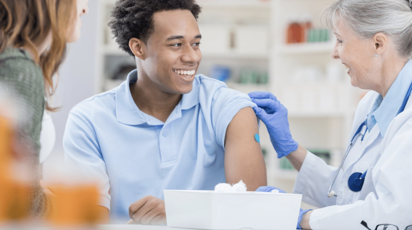 A smiling young man in a light blue shirt receives a vaccination from a healthcare professional wearing a white coat and blue gloves. A woman sits beside him. The setting appears to be a clinic or medical office with various supplies in the background.