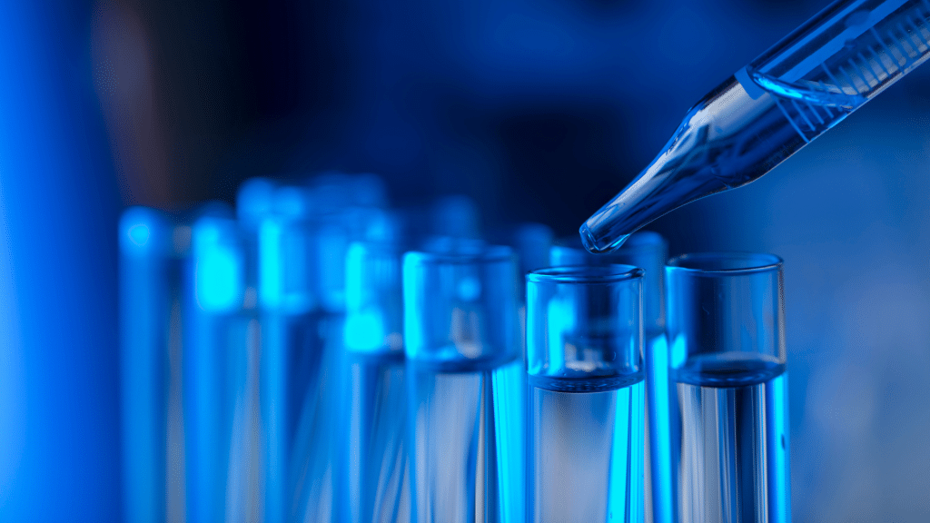 A series of transparent test tubes are arranged in rows with a liquid being dispensed into one of them using a precise dropper. The scene is illuminated with a blue hue, suggesting a controlled laboratory environment.