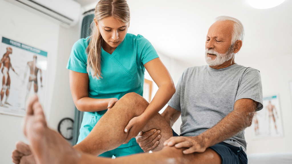 A healthcare worker in teal scrubs assists an elderly man with leg exercises in a bright room, likely a physical therapy setting. The man is seated and the worker supports his leg, focusing on therapy techniques.