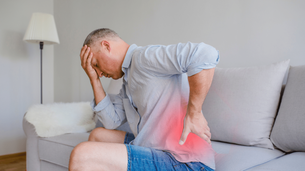 A man sitting on a couch, wearing a blue shirt and denim shorts, holds his lower back with one hand, while the other hand is on his forehead. He appears to be in pain, and there is a red area highlighted on his lower back to indicate discomfort.