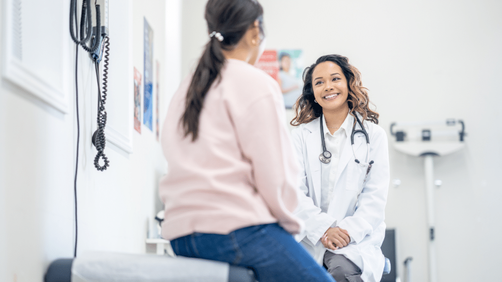 A doctor in a white coat sits and talks with a patient who is seated on an examination table. The patient is wearing a light pink sweater and jeans. The room contains medical equipment and a scale in the background.